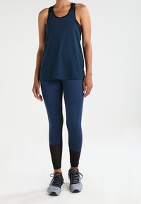 Canottiera blu navy con dettagli neri, abbinata a leggings blu navy con pannelli in rete e sezioni inferiori nere, completata da sneaker sportive grigie.