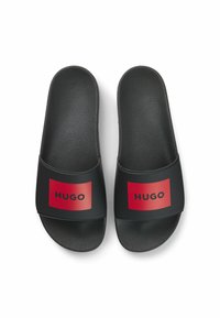 Infradito neri con soletta testurizzata, fasce larghe con un logo rettangolare rosso e lettere nere, suola piatta, design minimalista.
