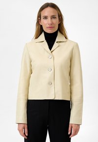Klippt beige läderjacka med notchad krage, med tre knappar och långa ärmar, tillsammans med en svart polotröja.