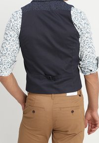 Marineblauwe vest met gestructureerde schouderdetails, verstelbare rugband, gedragen over een blauwe bloemenoverhemd, gecombineerd met lichtbruine broek.