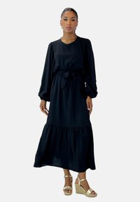 Schwarzes Langarmkleid mit einem gestuften Rock, Bindegürtel in der Taille und weichem Stoff. Kombiniert mit goldenen, strappy Wedge Sandalen.