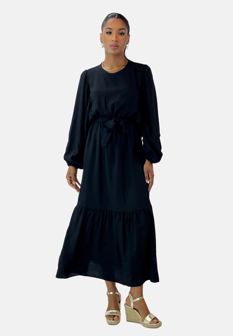 Schwarzes Langarmkleid mit einem gestuften Rock, Bindegürtel in der Taille und weichem Stoff. Kombiniert mit goldenen, strappy Wedge Sandalen.