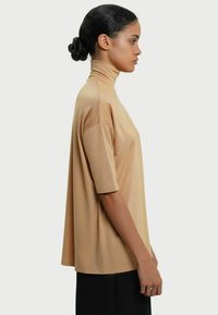 Beige højhalset kortærmet top, afslappet pasform, glat tekstur, blødt materiale, med en lang silhuet, vist på en model fra siden.