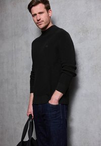 Pull en maille côtelée noir avec un col montant, comportant un détail de logo subtil. Associé à un jean en denim sombre et à un sac noir.
