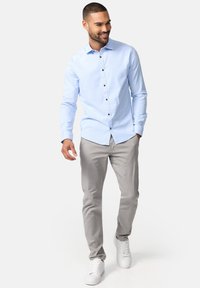 Chemise bleu clair à boutons marron, associée à un pantalon gris et des baskets blanches. La chemise présente une coupe ajustée et des manches longues.