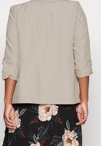 Blazer beige chiaro con maniche a tre quarti, texture liscia e vestibilità su misura. Indossato sopra una gonna nera con motivo floreale.