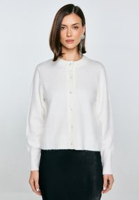 Bílý, nadýchaný kardigan s kulatým výstřihem, perlovými knoflíky vpředu a ribovanými manžetami, kombinovaný s černou sukní.