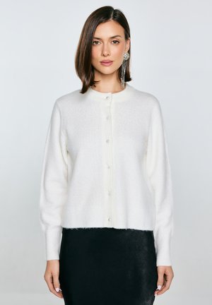 Biały, puszysty kardigan z okrągłym dekoltem, z perłowymi guzikami z przodu i ściągaczami na rękawach, zestawiony z czarną spódnicą.