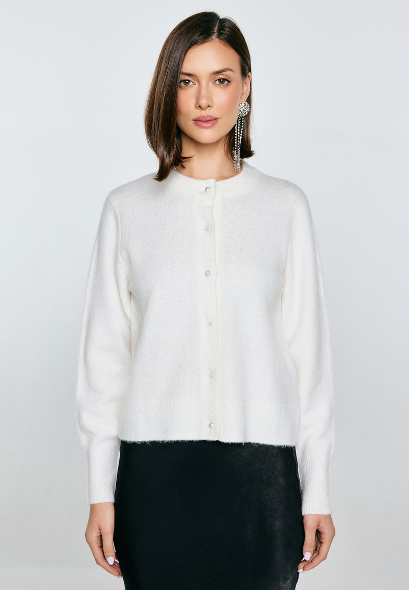Bílý, nadýchaný kardigan s kulatým výstřihem, perlovými knoflíky vpředu a ribovanými manžetami, kombinovaný s černou sukní.