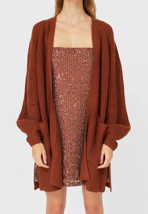 Femme portant un cardigan en maille épaisse couleur rouille par-dessus une mini-robe bustier ornée de sequins en or rose.