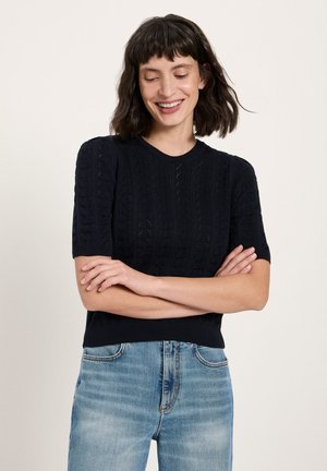 Lächelnde Frau mit kurzen dunklen Haaren, die einen schwarzen, strukturierten Kurzarmpullover und hellblaue Jeans trägt, mit verschränkten Armen.