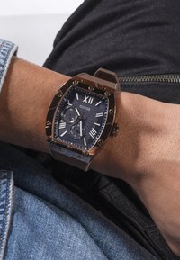 Rechteckige Uhr in Braun und Gold mit einem strukturierten dunkelblauen Zifferblatt, weißen römischen Ziffern, Unterzifferblättern und einem geflochtenen braunen Armband.