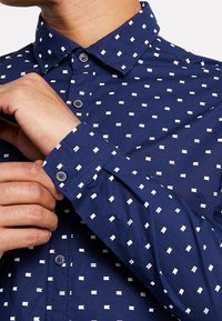 Chemise bleu marine à manches longues avec de petits motifs rectangulaires blancs. Présente un col classique et un poignet à boutons. Tissu doux et texturé.