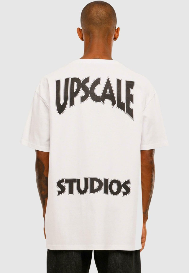 Biela bavlnená tričko s výrazným čiernym textom "UPSCALE" zakriveným hore a "STUDIOS" dole na zadnej strane. Voľný strih, krátke rukávy.