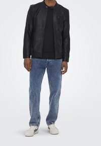 Uomo vestito con giacca di pelle nera, camicia nera, jeans blu e sneakers bianche, in piedi su uno sfondo chiaro uniforme.