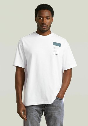 Mann trägt weißes oversized T-Shirt mit kleinem aufgedrucktem Label auf der linken Brust, kombiniert mit hellblauen Jeans, steht vor einfachem Hintergrund.