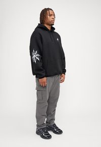 Felpa nera con un design di ragnatela sulla manica sinistra, abbinata a pantaloni cargo grigi e sneakers nere con accenti viola.