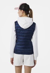 Mörkblå vadderad väst med quiltad textur, dragkedjestängning och en kontrasterande vit huvtröja under, bärs över vita shorts.
