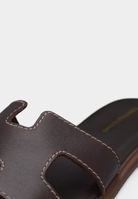 Κοντινό πλάνο καφέ δερμάτινου σανδαλιού σλάιντ με λευκή ραφή και διακοσμητικό κοψίμα στο πάνω λουράκι.