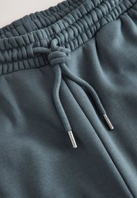 Gros plan d'une ceinture élastique gris foncé avec cordon noué et embouts métalliques sur un pantalon en tissu doux.