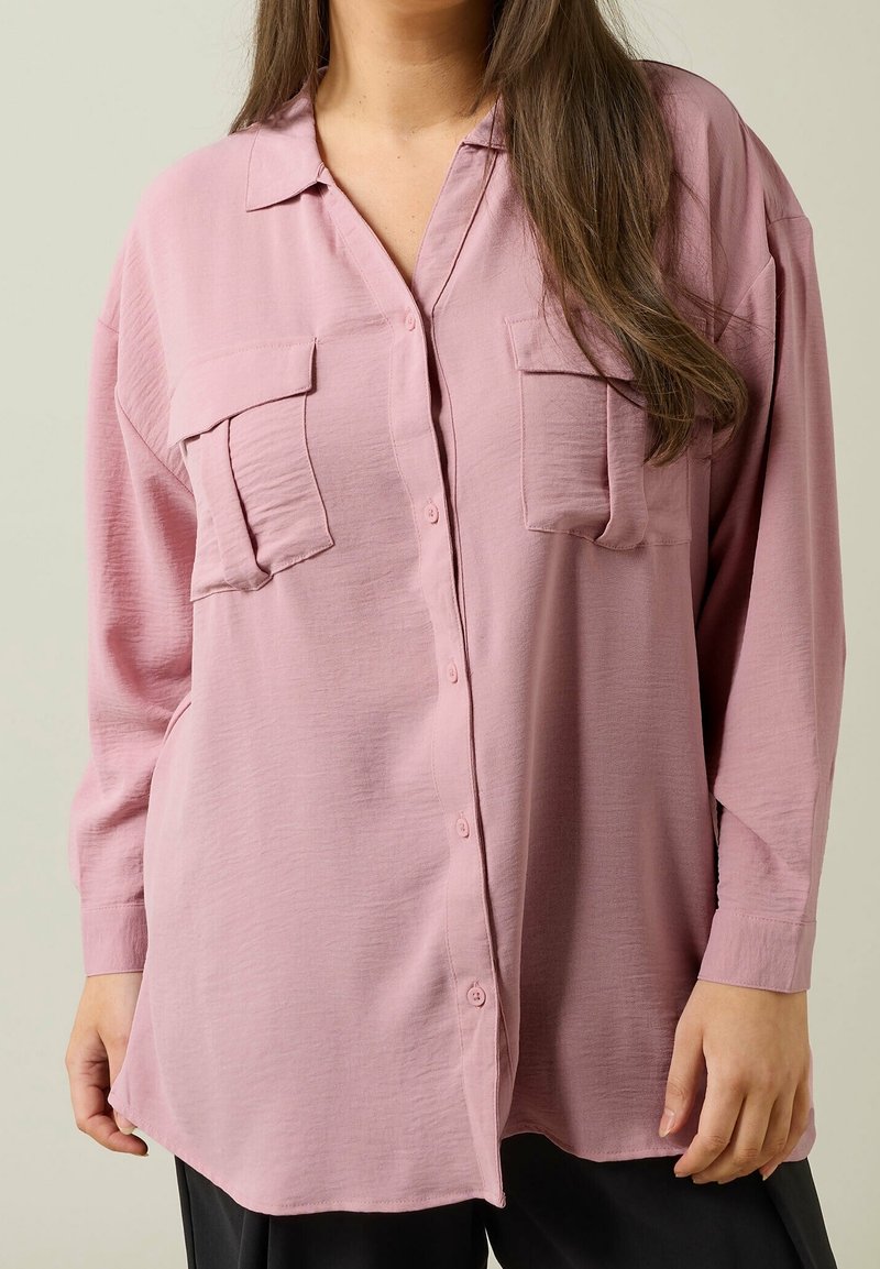 Femme portant une chemise rose ample à manches longues avec deux poches poitrine, montrée du cou aux hanches sur un fond uni.