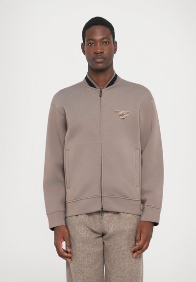 Sudadera gris con cremallera y cuello alzado, logotipo bordado en el lado izquierdo del pecho y bolsillos laterales; tela suave y ajuste relajado.