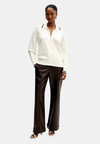 Maglione bianco con zip, dettagli neri e motivo floreale, abbinato a pantaloni in ecopelle marrone. Il modello indossa sandali con apertura e cinturini.