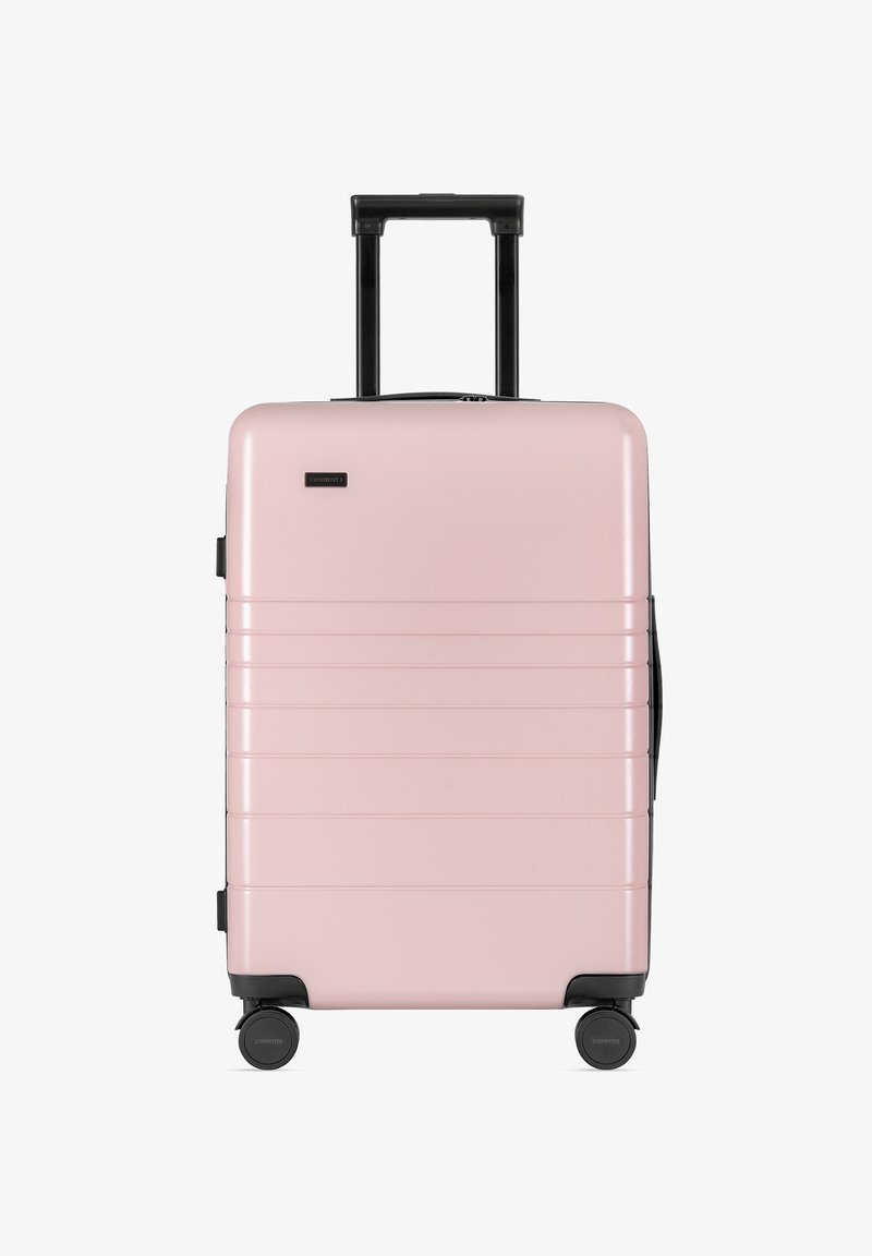 Hellrosa Hartschalenkoffer mit schwarzem Griff und Rädern, ausgestattet mit horizontalen Rillen und einem kleinen Logo auf der Vorderseite. Kompaktes Design.