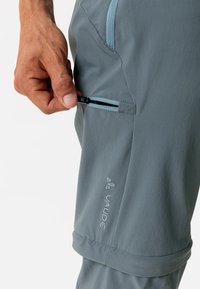 Lichtgrijze wandelbroeken met een gladde textuur, voorzien van een ritsvak en geborduurd logo-detail. Accentstiksels in een contrasterend blauw.