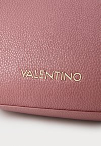 Rožinė tekstūruota odinė rankinė, papuošta auksiniu „VALENTINO“ logotipu. Paviršius turi akmenėtą tekstūrą ir yra sukurtas su švariomis linijomis.