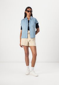 Lyseblå denim kortermet skjorte lagd over en marineblå t-skjorte med grafikk, kombinert med beige skreddersydde shorts og hvite joggesko.