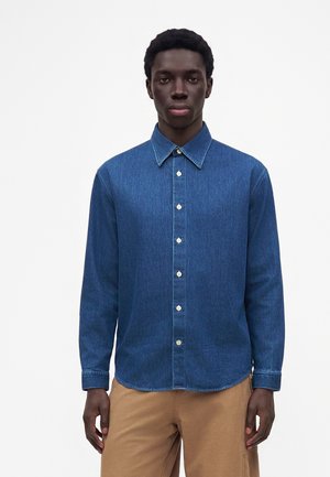 Homme debout face à l'avant, portant une chemise en jean bleue à boutons et un pantalon marron clair sur un fond blanc uni.