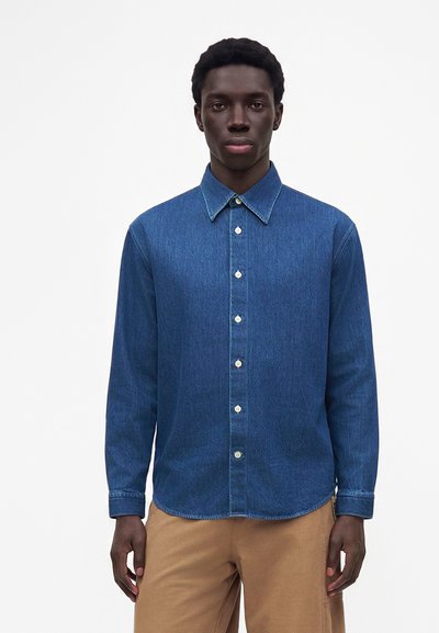 Homme debout face à l'avant, portant une chemise en jean bleue à boutons et un pantalon marron clair sur un fond blanc uni.