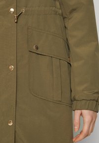 Olivgrüne Parka mit goldfarbenen Beschlägen, ausgestattet mit einer Brusttasche, elastischen Bündchen und einem Reißverschluss. Glatte Textur mit Nahtdetails.