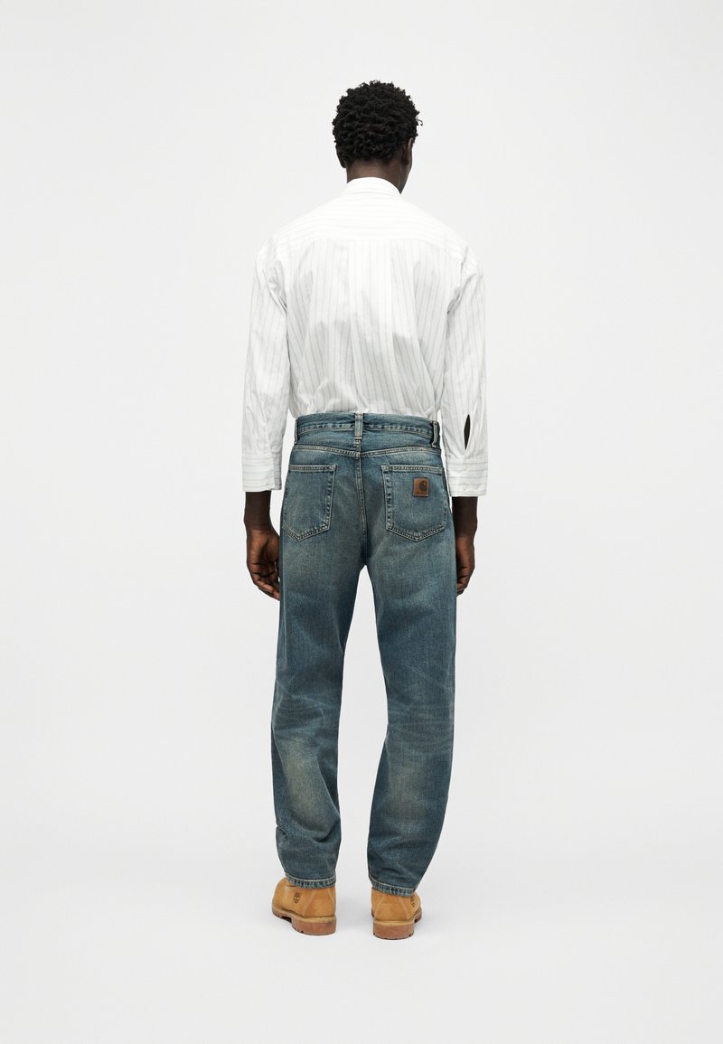 Homme debout, dos tourné, portant un jean denim bleu clair, une chemise blanche à rayures et des bottes de travail beige sur un fond uni.