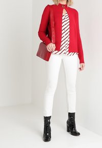 Roter gerippter Cardigan mit Knopfdetails, gestreifte Bluse in Weiß und Schwarz, weiße Hose und glänzende schwarze Stiefeletten, eine rote Tasche tragend.