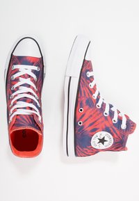 Zapatillas Converse de caña alta con un patrón de tie-dye en rojo y azul marino, punteras de goma blanca, cordones blancos y logo de estrella en el tobillo.