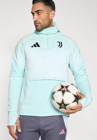 Lyseblå sports-pullover med halv lynlås, pink accenter og sorte logoer; kombineret med grå bukser og holder en mønstret fodbold.