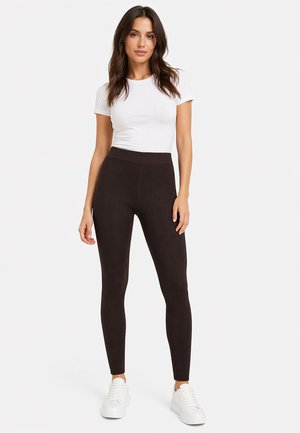 Frau steht, trägt ein weißes Kurzarmoberteil, schwarze gerippte Leggings mit hoher Taille und weiße Turnschuhe vor einem einfarbigen Hintergrund.