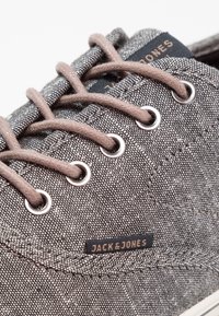 Zapatillas de tela grises con cordones marrones, ojales de metal y una etiqueta de "JACK & JONES". Superficie texturizada con un patrón sutil.