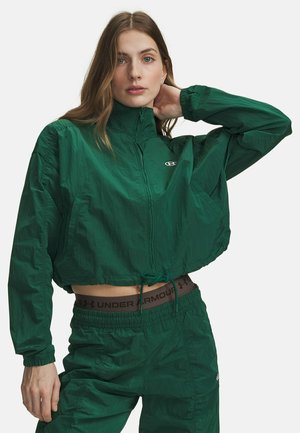 Jonge vrouw draagt een groene cropped jas met ritssluiting en bijpassende broek met elastische tailleband en trekkoord, poseert met hand achter de nek.