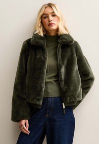 Chaqueta de piel sintética verde oliva con cuello de solapa, cierre de cremallera y un corte holgado, usada sobre una camiseta verde acanalada y vaqueros azul oscuro.