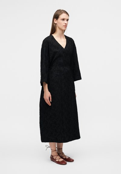 Robe longue noire portefeuille avec un motif floral texturé, décolleté en V et manches trois-quarts, associée à des chaussures plates marron à lacets.