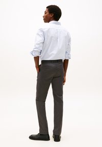 Homem de pé com as mãos nos bolsos, vestindo camisa azul clara às riscas, calças cinza escuro e sapatos pretos, de perfil sobre fundo branco.