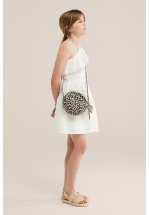 Ragazza giovane con vestito bianco monospalla, borsa a tracolla rotonda con stampa leopardata e sandali beige, in piedi di profilo su sfondo neutro.