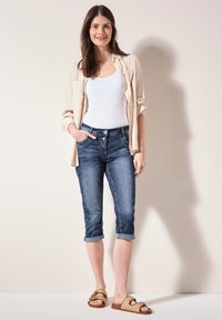 Camisa de botón beige clara sobre un top blanco sin mangas, combinada con jeans oscuros recortados y sandalias de tiras trenzadas en color tostado. Texturas suaves y puños enrollados.