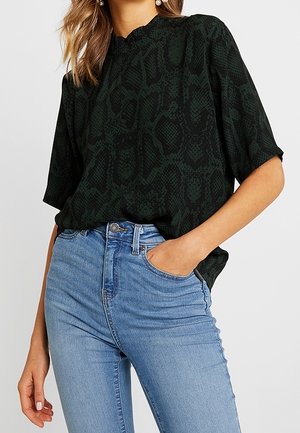 Frau trägt eine dunkelgrüne Bluse mit Schlangenprint und kurzen Ärmeln, in eine hoch taillierte hellblaue Jeans gesteckt, mit einer Hand in der Tasche.