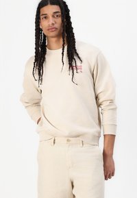 Beige sweatshirt with ribbed cuffs and hem, featuring red text graphic on chest. Worn with light beige pants, displaying a smooth texture.