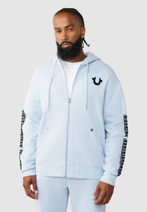 Mann mit geflochtenem Haar und Bart, trägt hellblauen Hoodie mit Reißverschluss und passende Hose, steht vor schlichtem grauem Hintergrund.