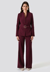 Bordeaux kleurige op maat gemaakte pantsuit met een verstelbare taille, scherpe revers en brede pijpen. Gladde stof en minimalistisch ontwerp.